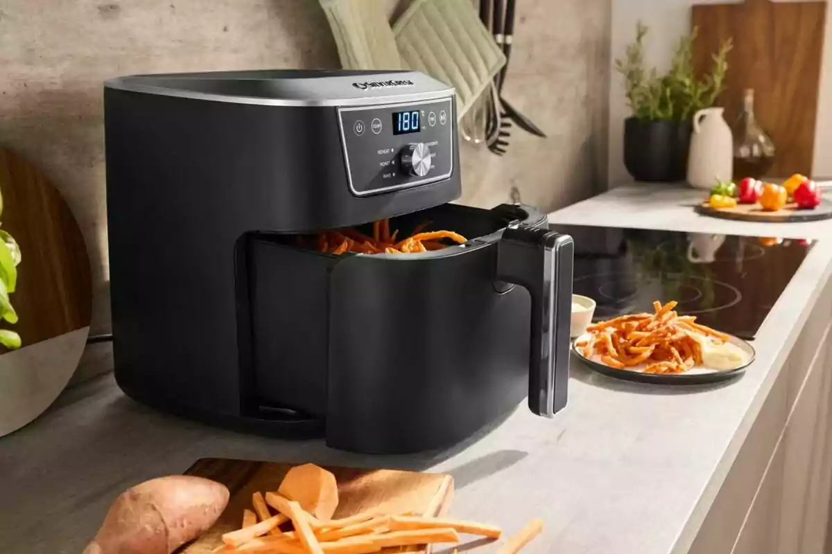 Freidora de aire negra cocinando papas fritas en una cocina moderna con ingredientes frescos y utensilios al fondo Freidora de aire negra cocinando papas fritas en una cocina moderna con ingredientes frescos y utensilios al fondo