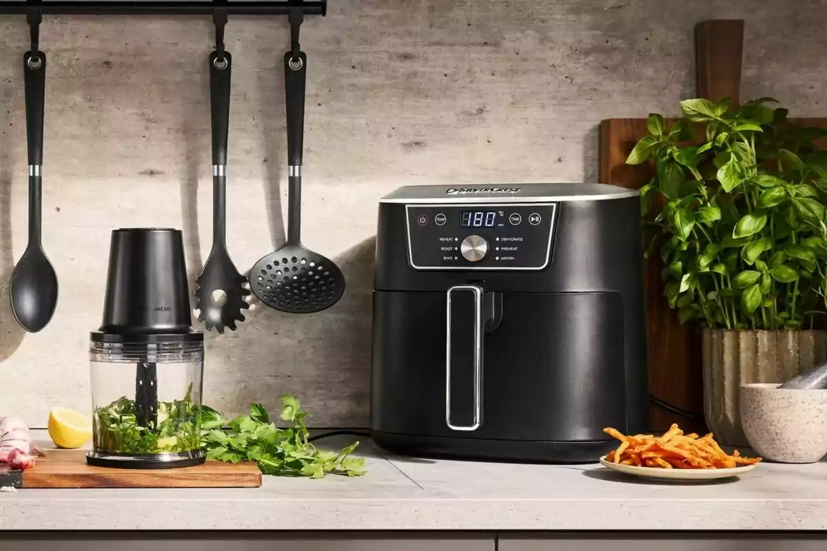 Freidora de aire negra sobre una encimera de cocina junto a un procesador de alimentos, utensilios colgados, un plato con papas fritas y una planta en maceta Freidora de aire negra sobre una encimera de cocina junto a un procesador de alimentos, utensilios colgados, un plato con papas fritas y una planta en maceta