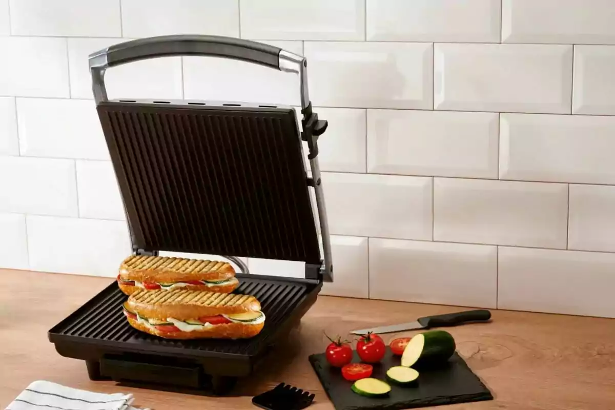 Sandwichera eléctrica abierta con dos paninis y vegetales frescos sobre una mesa de cocina