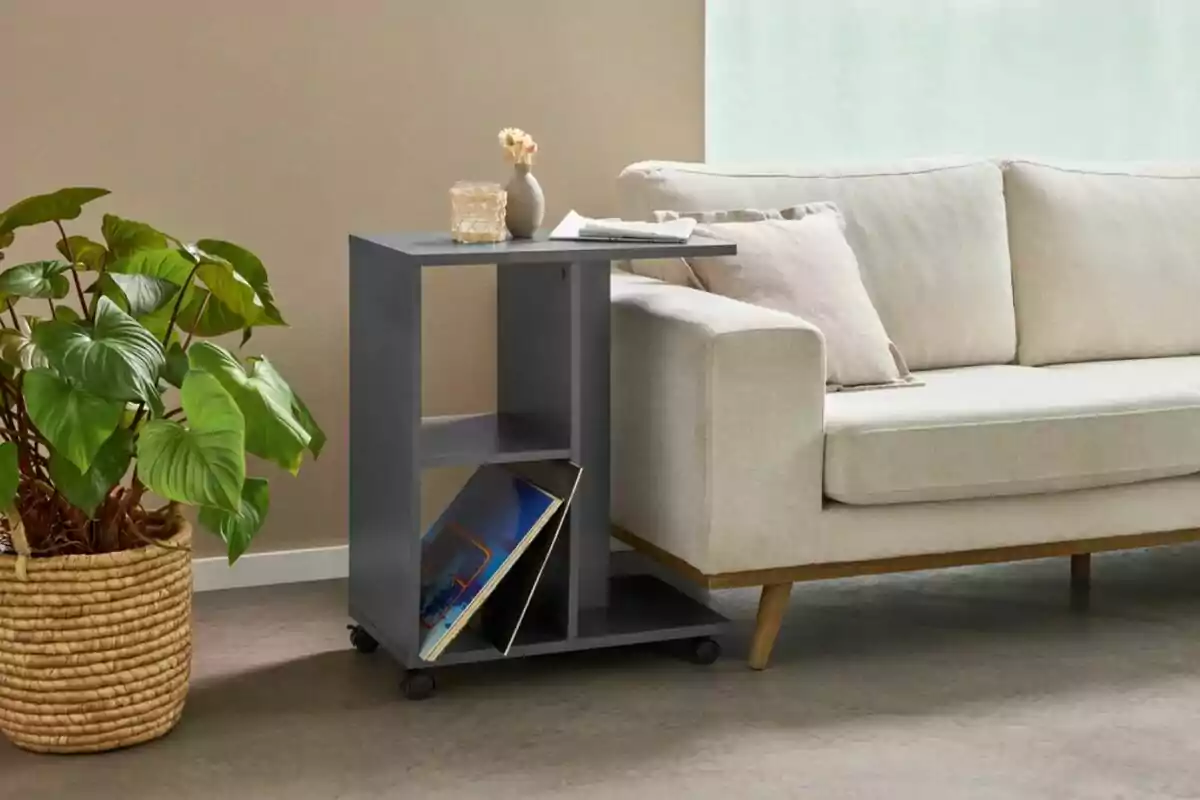Mesa auxiliar gris con ruedas junto a un sofá beige, con una planta en maceta de mimbre al lado y algunos objetos decorativos y revistas sobre la mesa Mesa auxiliar gris con ruedas junto a un sofá beige, con una planta en maceta de mimbre al lado y algunos objetos decorativos y revistas sobre la mesa