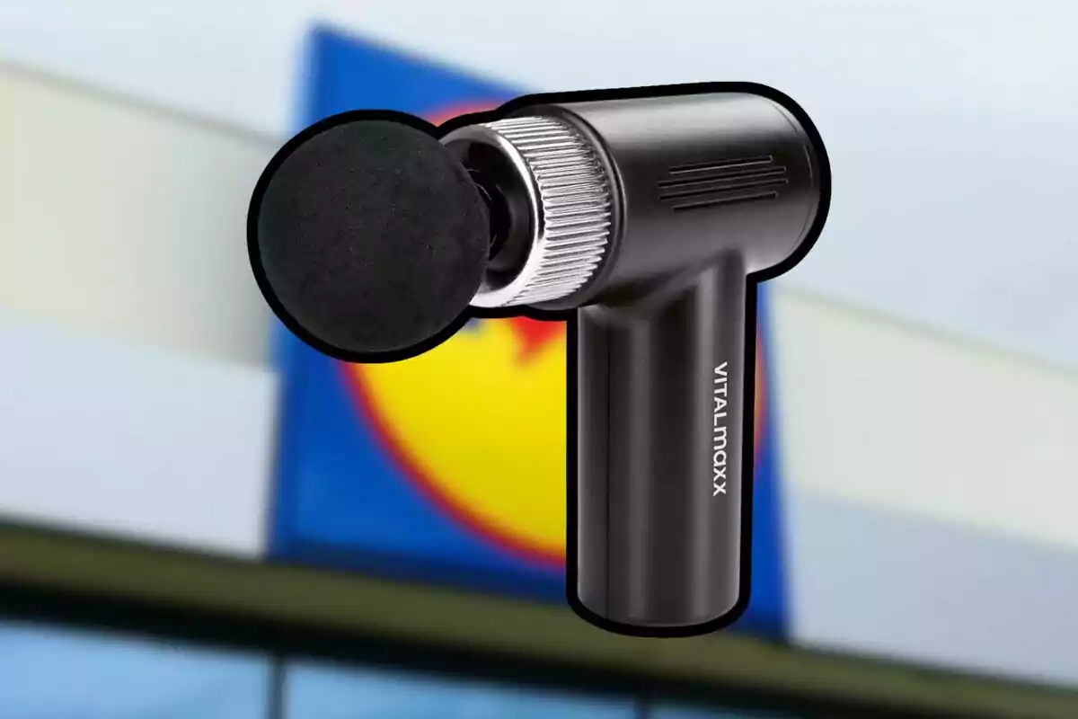 Pistola de masaje negra de la marca Vitalmaxx sobre un fondo desenfocado de un letrero de supermercado.