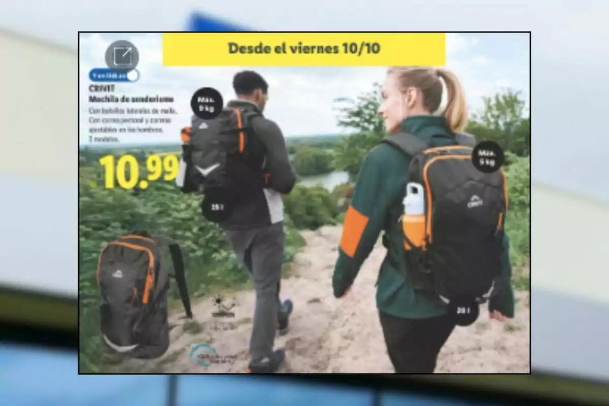 Dos personas caminan por un sendero con mochilas negras y detalles naranjas mientras un anuncio promociona mochilas de senderismo Crivit por 10.99 euros disponibles desde el viernes 10 de octubre