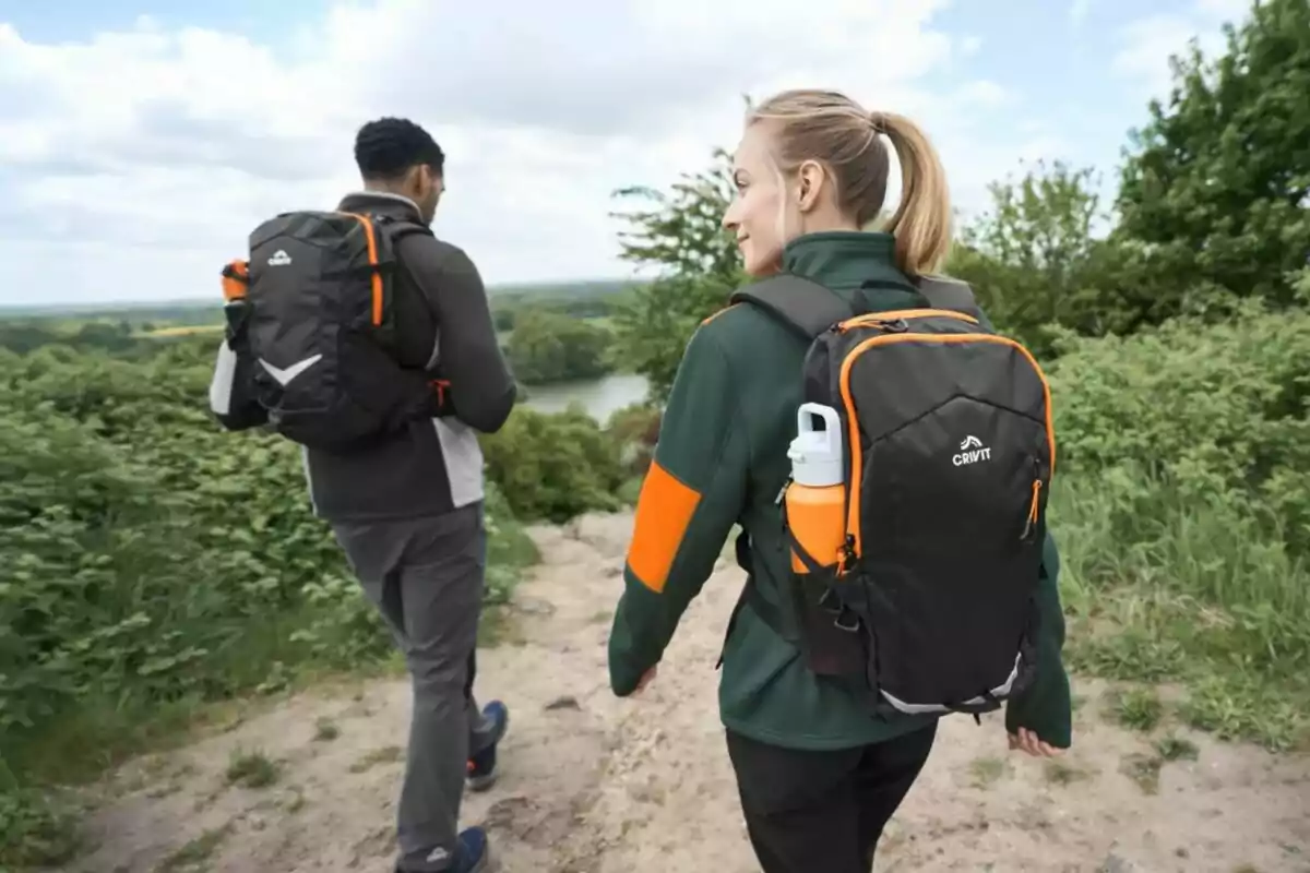Dos personas con mochilas de senderismo caminan por un sendero rodeado de vegetación y árboles, con un lago visible al fondo