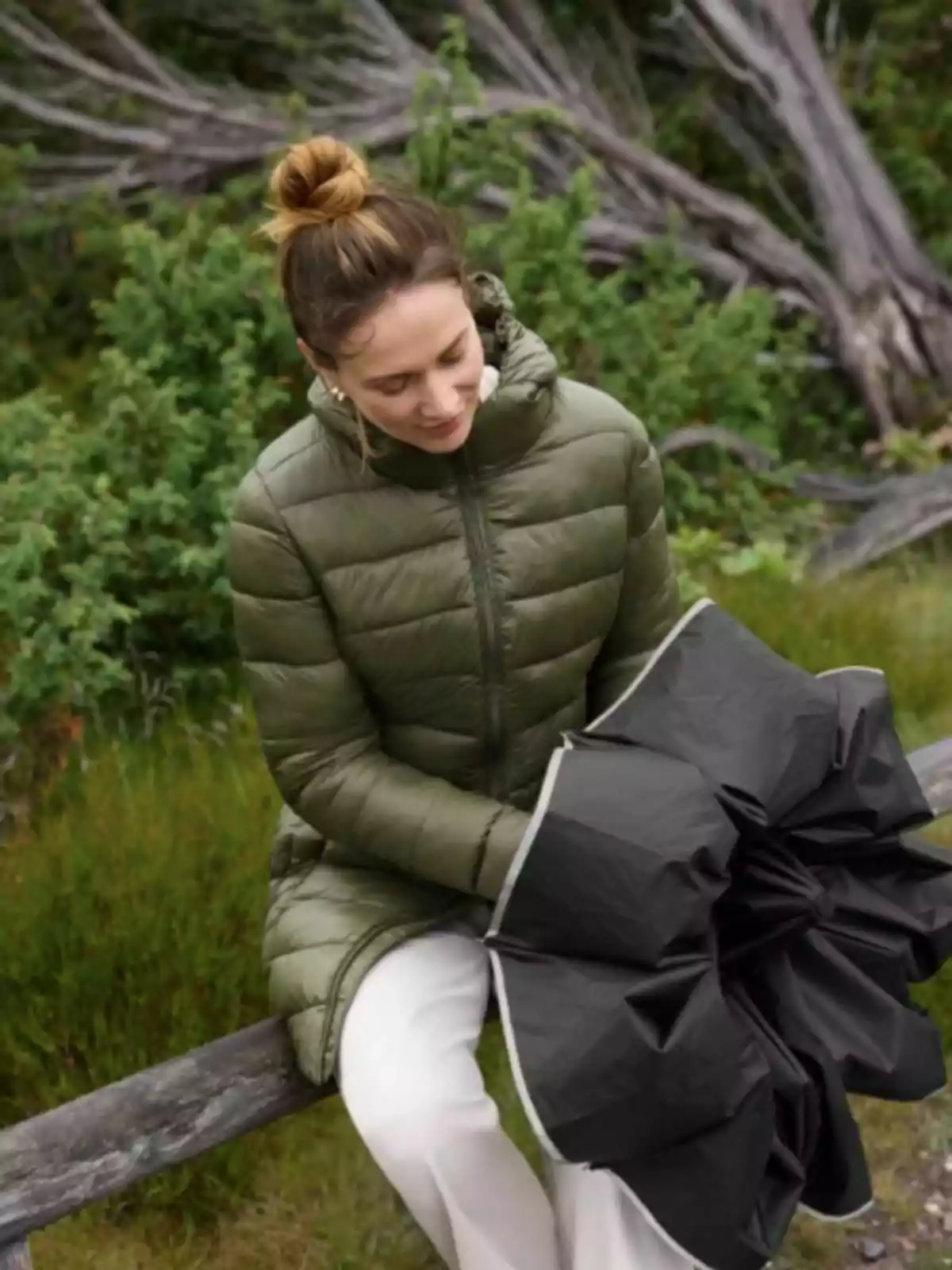 Dona amb abric verd i pantalons clars asseguda en una tanca de fusta sostenint un paraigua negre tancat en un entorn natural