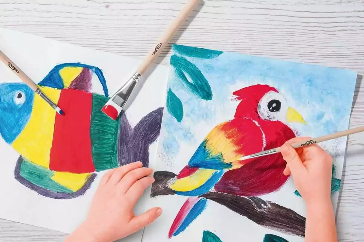 Dos manos de niño pintando un pez y un loro coloridos con pinceles sobre hojas de papel blanco