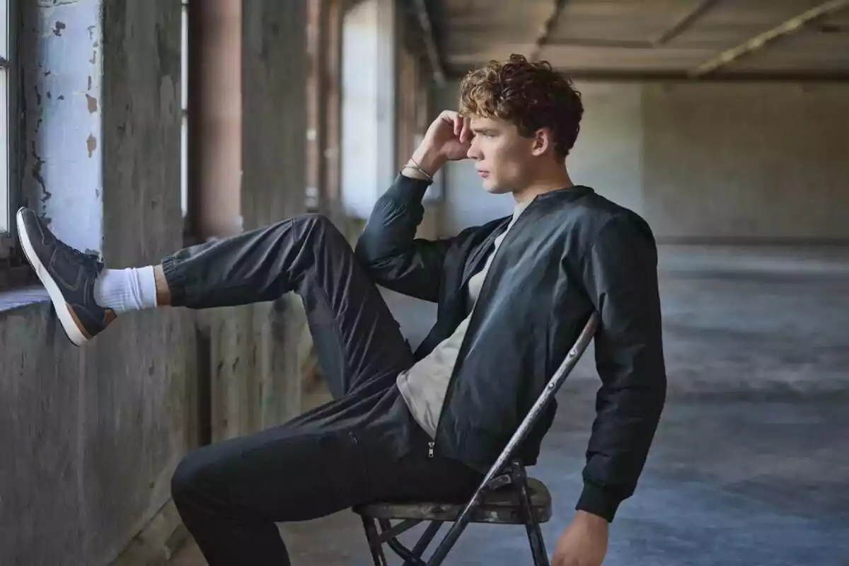 Joven sentado en una silla metálica dentro de un edificio industrial con la pierna apoyada en la ventana y mirando pensativo hacia afuera Joven sentado en una silla metálica dentro de un edificio industrial con la pierna apoyada en la ventana y mirando pensativo hacia afuera