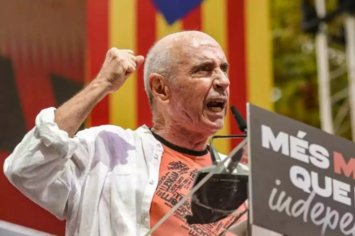 Home gran parlant apassionadament en un míting amb el puny alçat i una pancarta davant seu amb un fons de bandera catalana