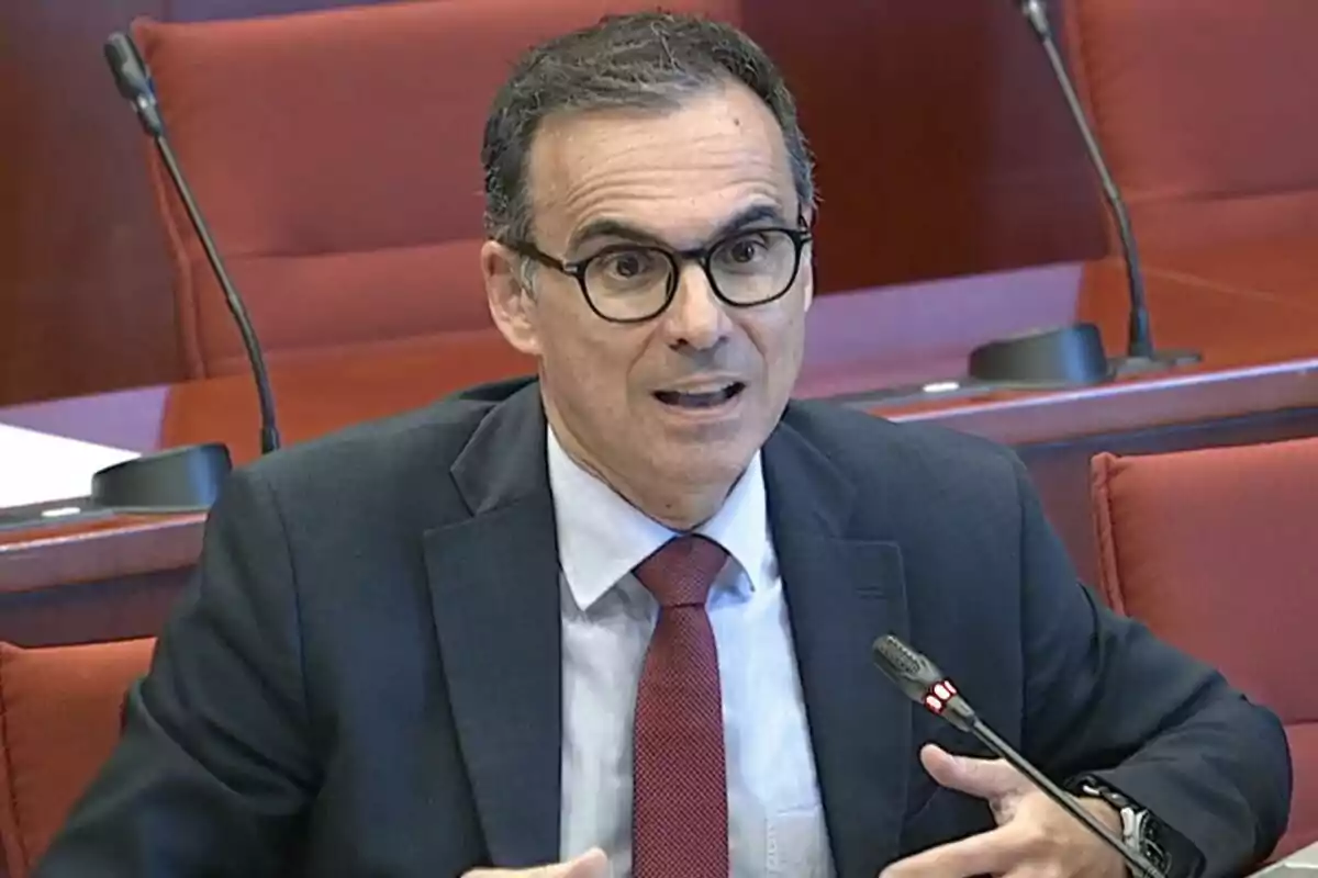 Hombre con gafas y traje oscuro hablando frente a un micrófono en una sala de conferencias con asientos rojos