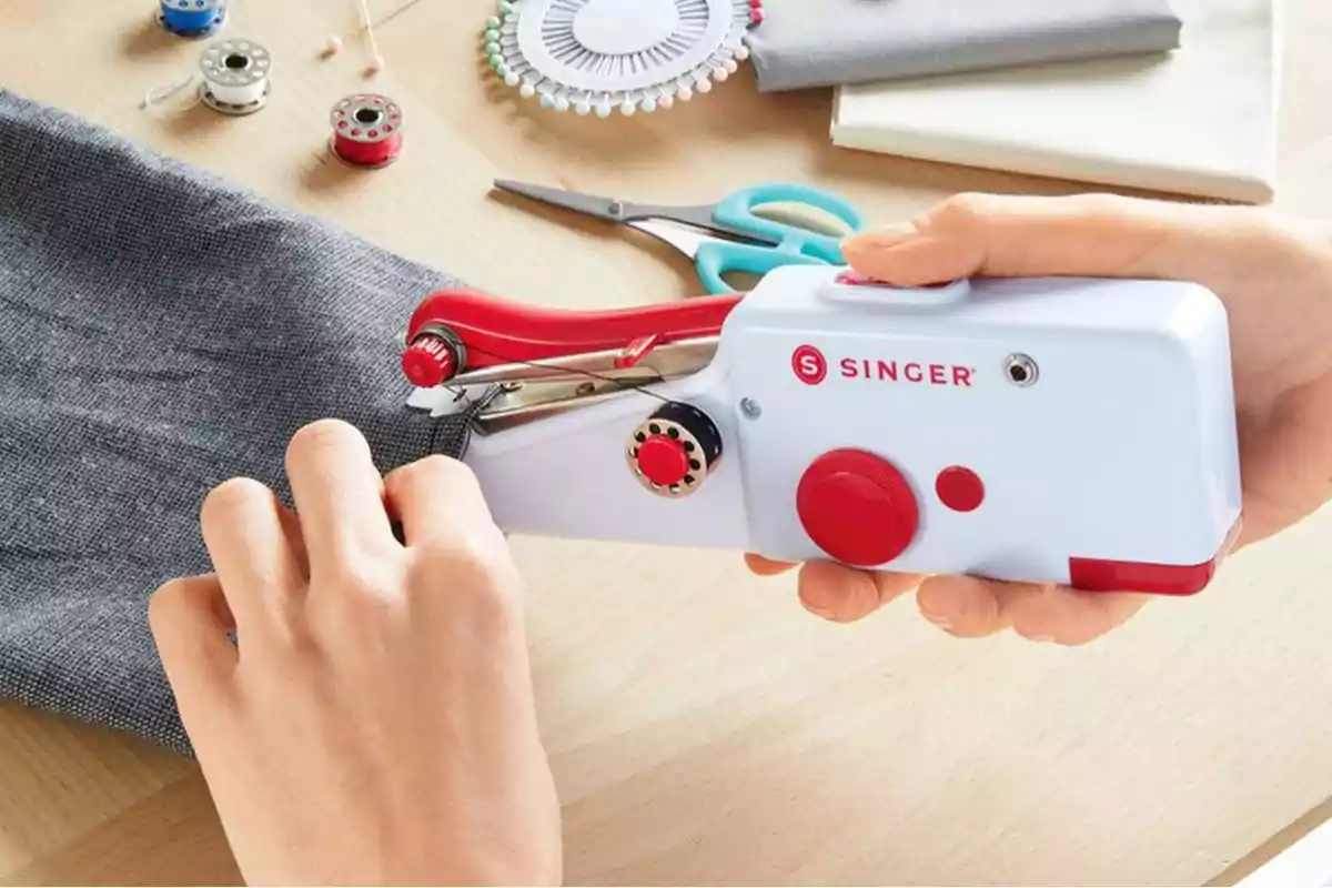 Person using a portable Singer sewing machine to sew gray fabric on a table with sewing accessories around.