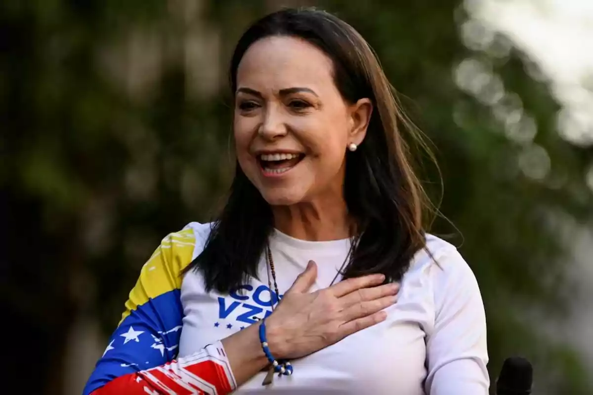 Mujer sonriente con el cabello oscuro y una camiseta con los colores de la bandera de Venezuela, con la mano en el pecho y fondo desenfocado de árboles.