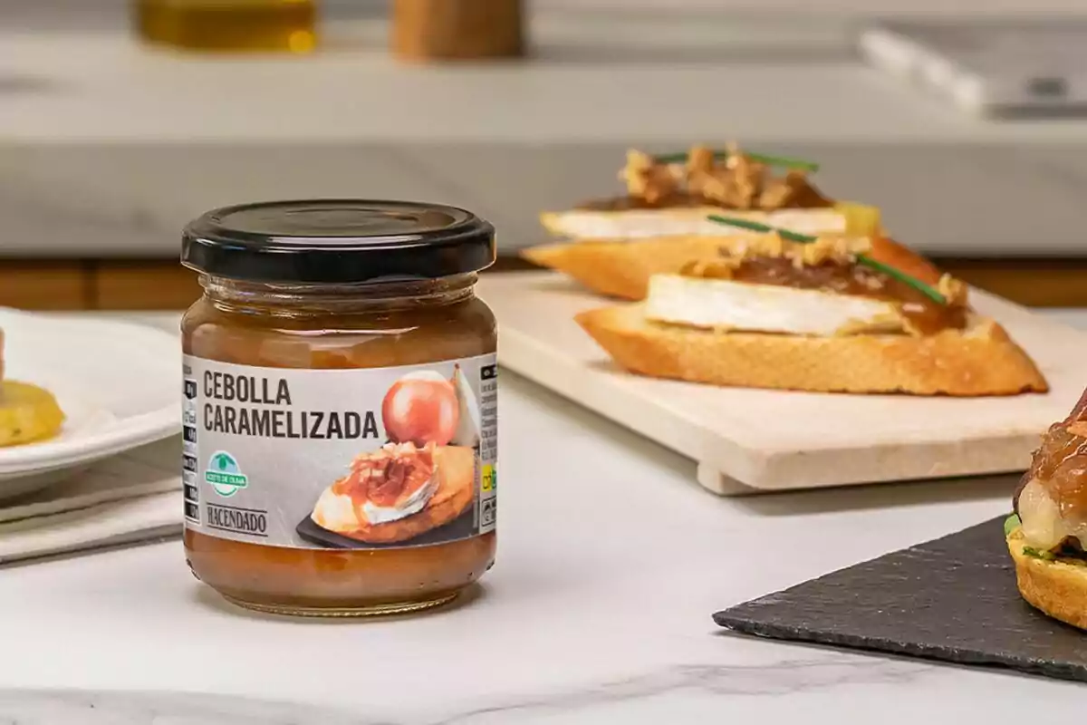 Un frasco de cebolla caramelizada sobre una mesa junto a unas tostadas con queso y cebolla caramelizada como aperitivo