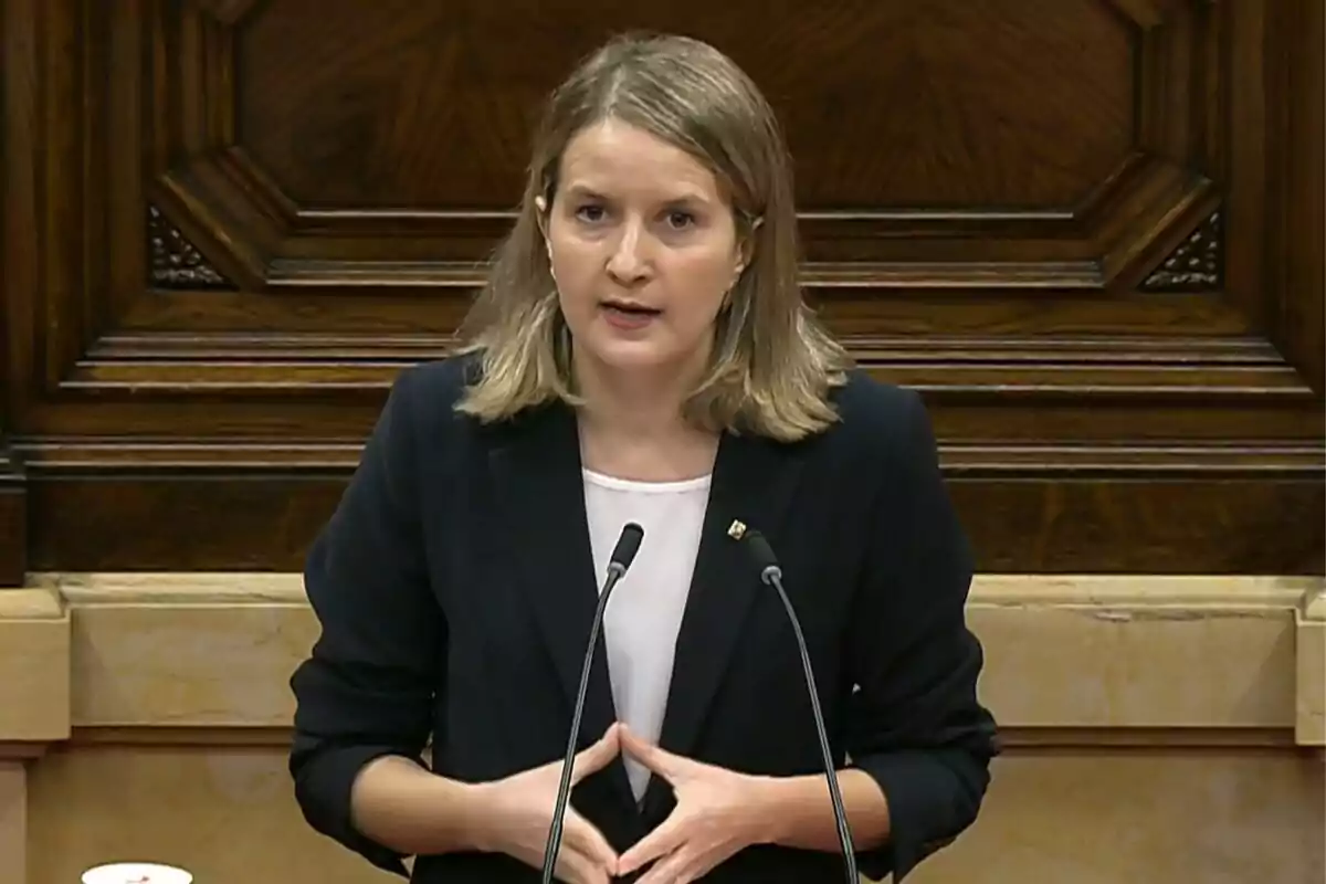 Mujer de cabello rubio y liso hablando en un podio con fondo de madera y dos micrófonos frente a ella
