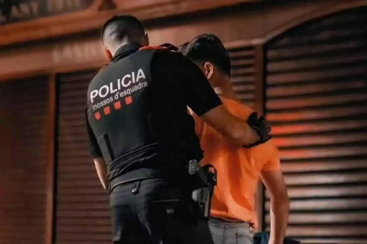 Un agent dels Mossos d'Esquadra fa un control a un home amb samarreta taronja davant d'una persiana tancada