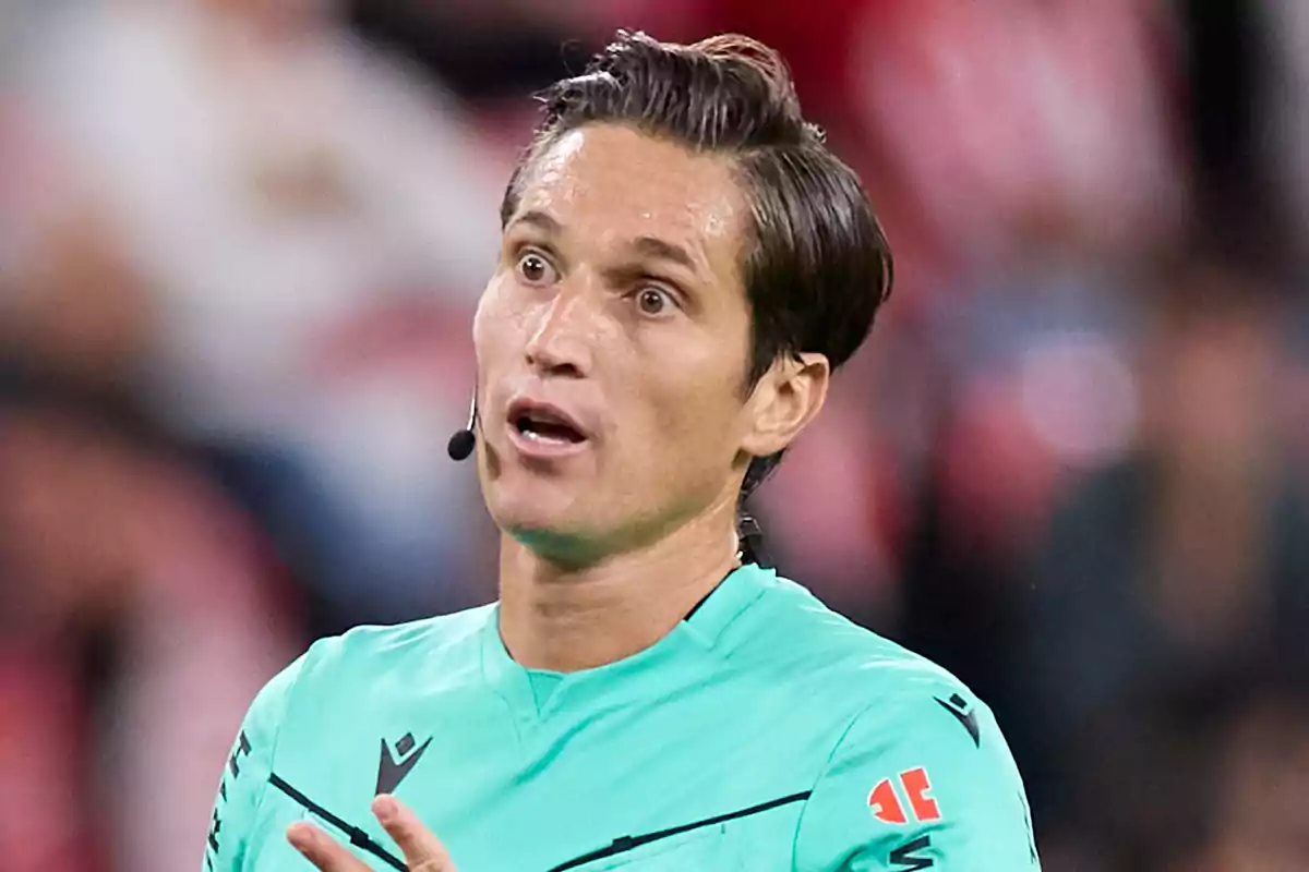 Árbitro de fútbol con uniforme verde y micrófono en la oreja gesticulando durante un partido