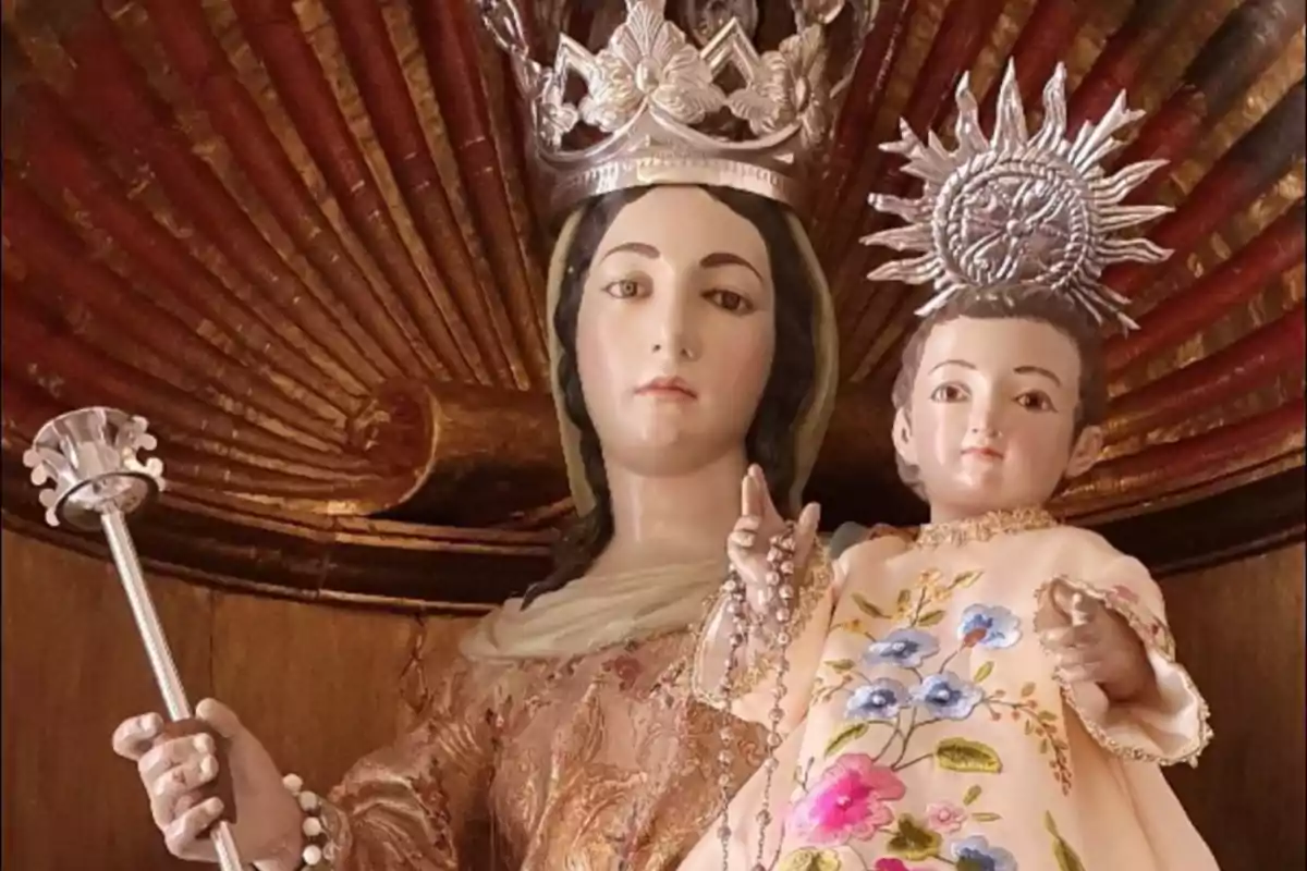 Imagen de una estatua religiosa de la Virgen María con corona sosteniendo al Niño Jesús, ambos vestidos con ropas decoradas y detalles metálicos en la cabeza, fondo de madera tallada.