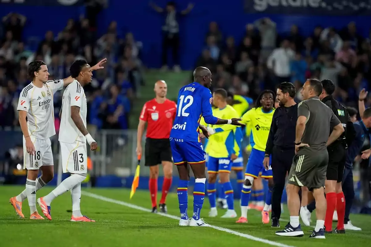 Jugadores de dos equipos de fútbol discuten en la banda mientras el árbitro observa y los entrenadores intervienen