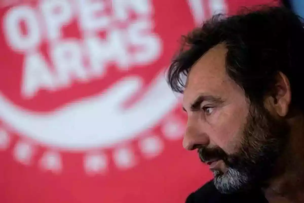 Hombre de perfil con barba frente a un fondo rojo con el logo de Open Arms desenfocado