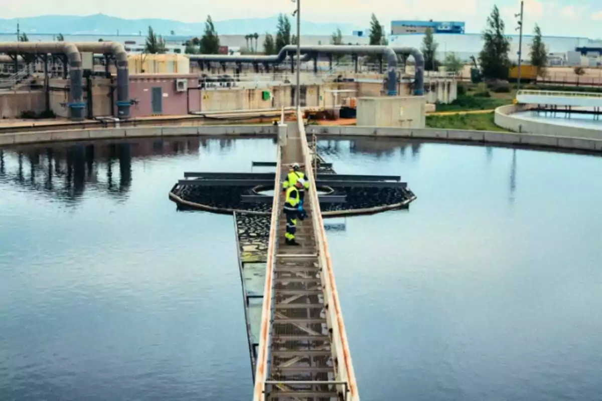 Treballador amb armilla de seguretat caminant per una passarel·la en una planta de tractament d’aigües envoltada de grans dipòsits circulars i canonades industrials.