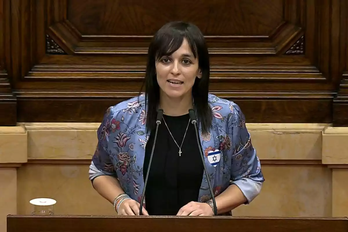 Dona de cabells foscos parlant en un faristol de fusta amb un fons de panells de fusta, porta una jaqueta blava amb flors i una insígnia amb la bandera d’Israel. Dona de cabells foscos parlant en un faristol de fusta amb un fons de panells de fusta, porta una jaqueta blava amb flors i una insígnia amb la bandera d’Israel.