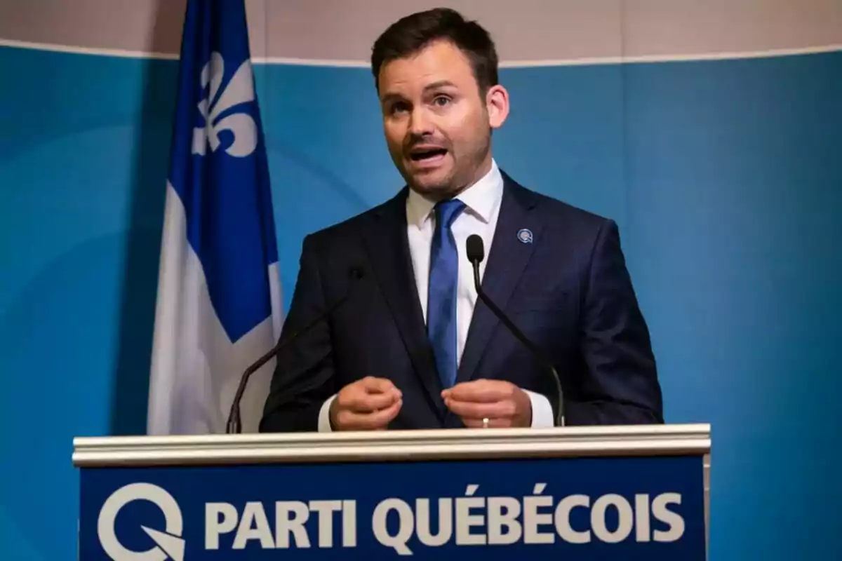 Hombre con traje azul hablando en un podio del Parti Québécois con una bandera azul y blanca de fondo