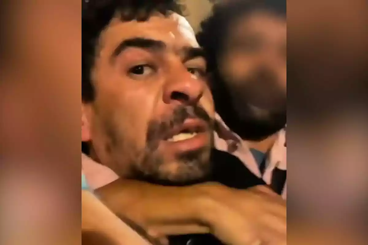 Hombre con expresión de angustia siendo sujetado por otra persona en un entorno poco iluminado