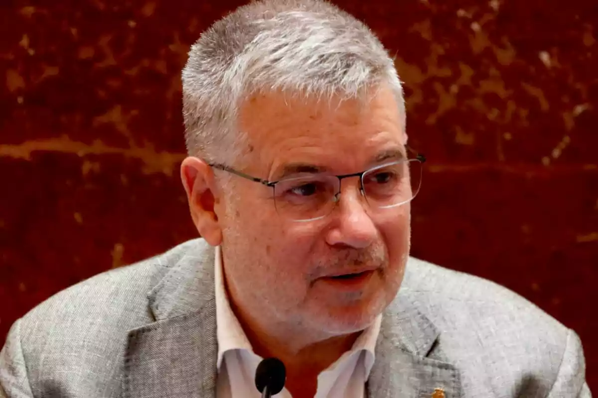 Hombre de cabello canoso y gafas hablando frente a un micrófono con fondo marrón