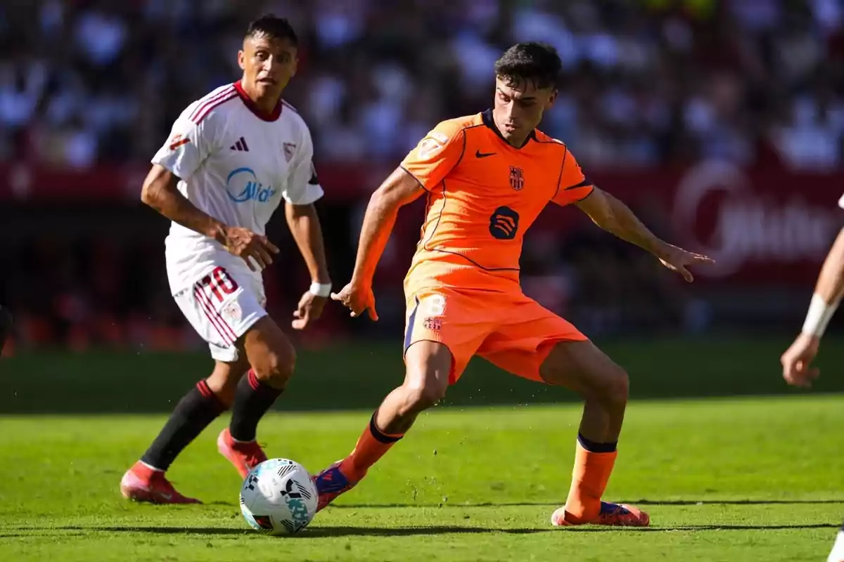 Dos futbolistes disputen la pilota durant un partit en un estadi ple d’espectadors; un porta uniforme blanc i l’altre uniforme taronja.