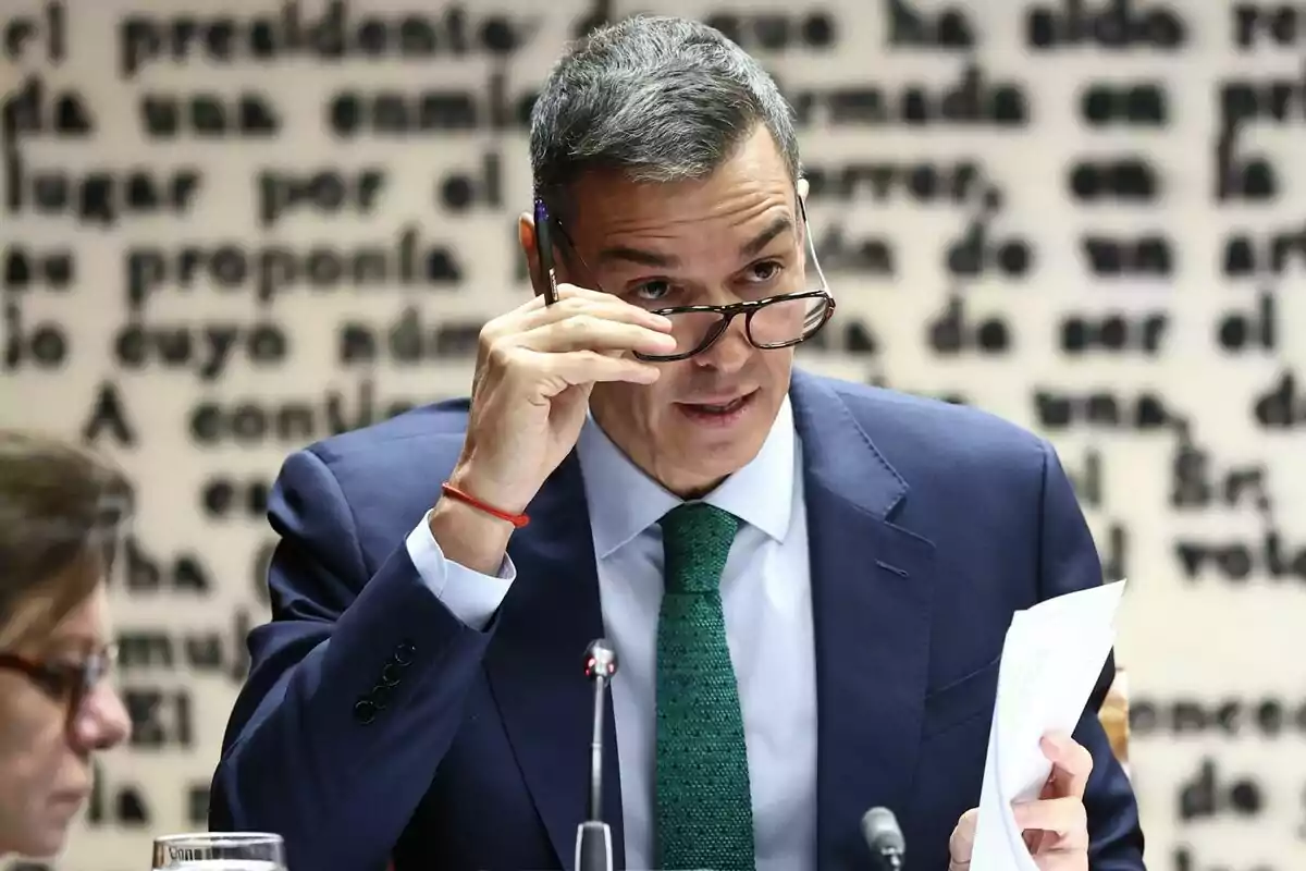 Hombre de traje azul y corbata verde ajustándose las gafas mientras sostiene unos papeles en una reunión con fondo de letras desenfocadas Hombre de traje azul y corbata verde ajustándose las gafas mientras sostiene unos papeles en una reunión con fondo de letras desenfocadas