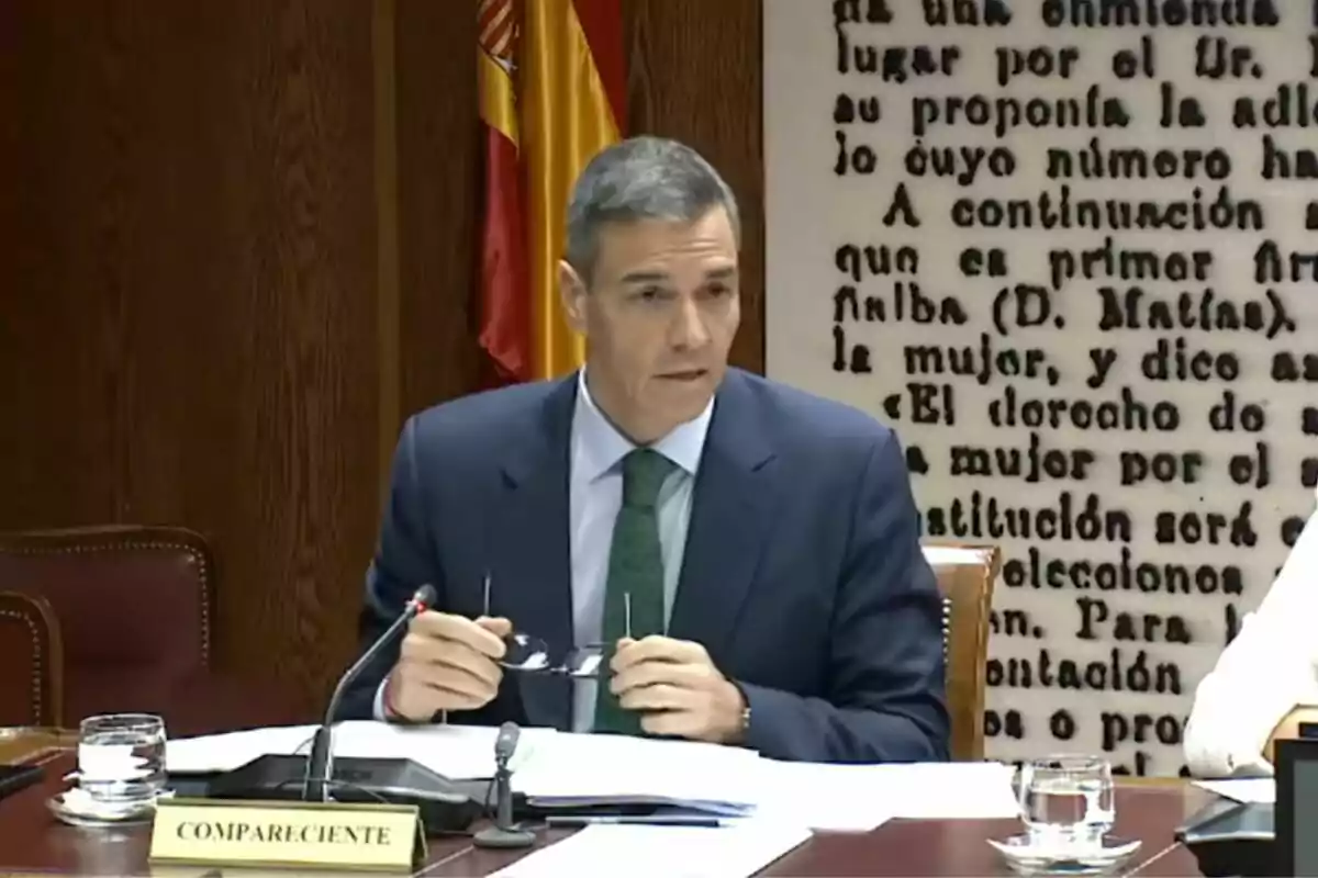 Hombre de traje sentado en una mesa de reunión con documentos y micrófono frente a él, bandera de España y texto en la pared de fondo