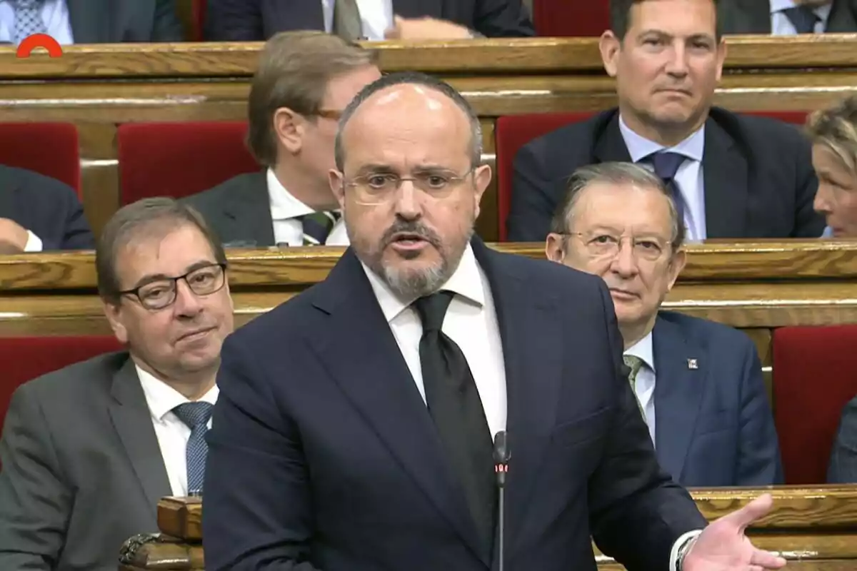 Un hombre de traje y corbata habla en un parlamento mientras varias personas lo observan desde sus asientos Un hombre de traje y corbata habla en un parlamento mientras varias personas lo observan desde sus asientos
