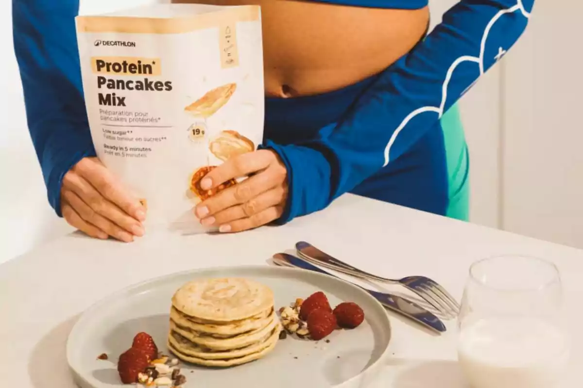 Persona amb roba esportiva sosté un paquet de barreja per a pancakes de proteïna de Decathlon al costat d’un plat amb pancakes, gerds, nous i un got de llet.