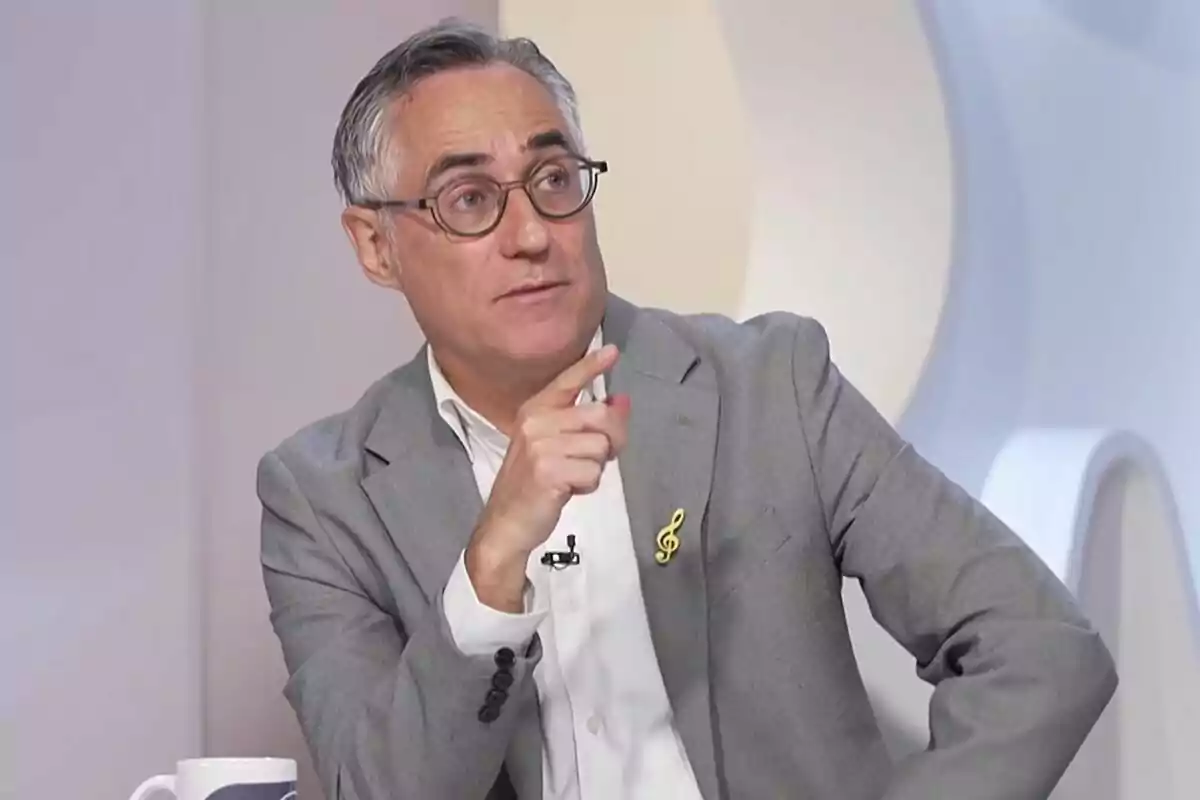Hombre de cabello canoso y gafas con saco gris y camisa blanca señalando con el dedo mientras está sentado en un estudio de televisión