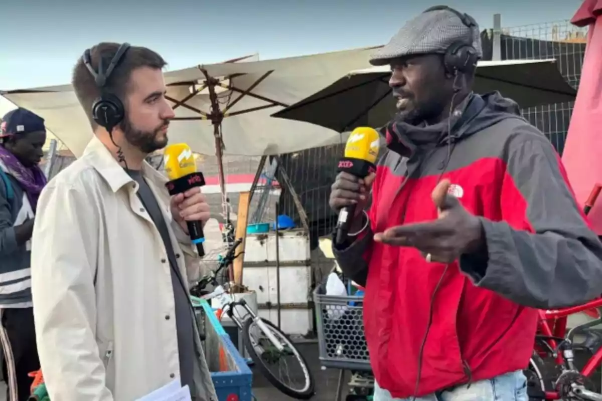 Dos hombres con auriculares y micrófonos conversan al aire libre, rodeados de bicicletas y sombrillas, mientras uno de ellos gesticula con la mano. Dos hombres con auriculares y micrófonos conversan al aire libre, rodeados de bicicletas y sombrillas, mientras uno de ellos gesticula con la mano.