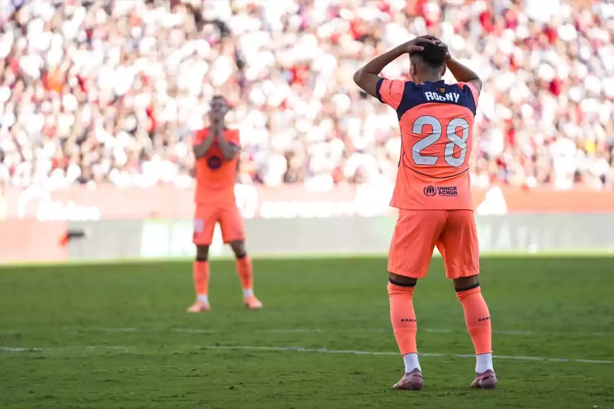 Dos futbolistas con uniforme naranja muestran gestos de frustración en el campo durante un partido, uno con las manos en la cabeza y el otro al fondo con las manos en la cara, mientras el público observa desde las gradas.