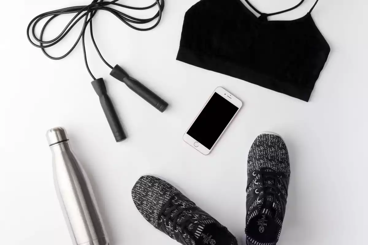 Ropa deportiva negra, un par de tenis, una cuerda para saltar, una botella de agua metálica y un teléfono móvil sobre una superficie blanca
