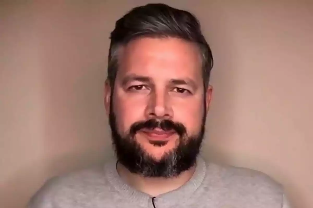 Home de cabell curt i barba fosca amb jersei gris posant davant d’un fons beix