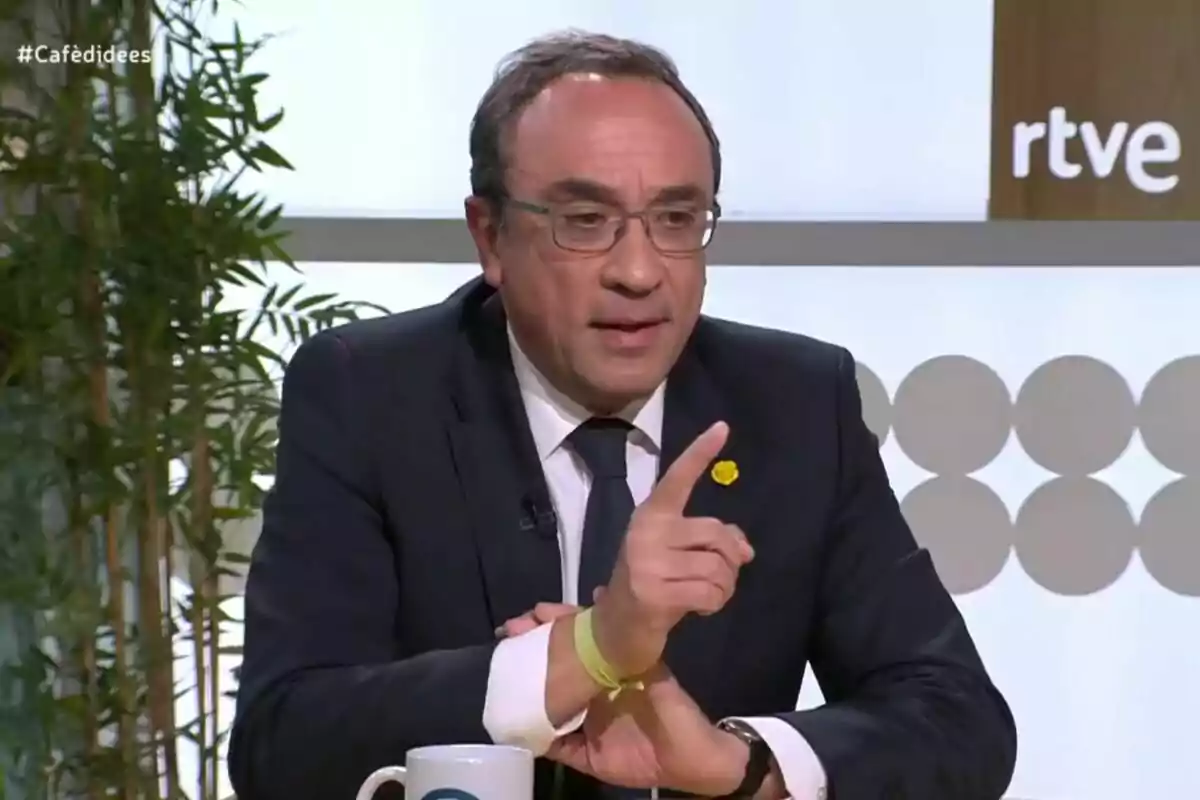 Hombre con gafas y traje oscuro hablando en un programa de televisión con una taza blanca sobre la mesa y plantas al fondo