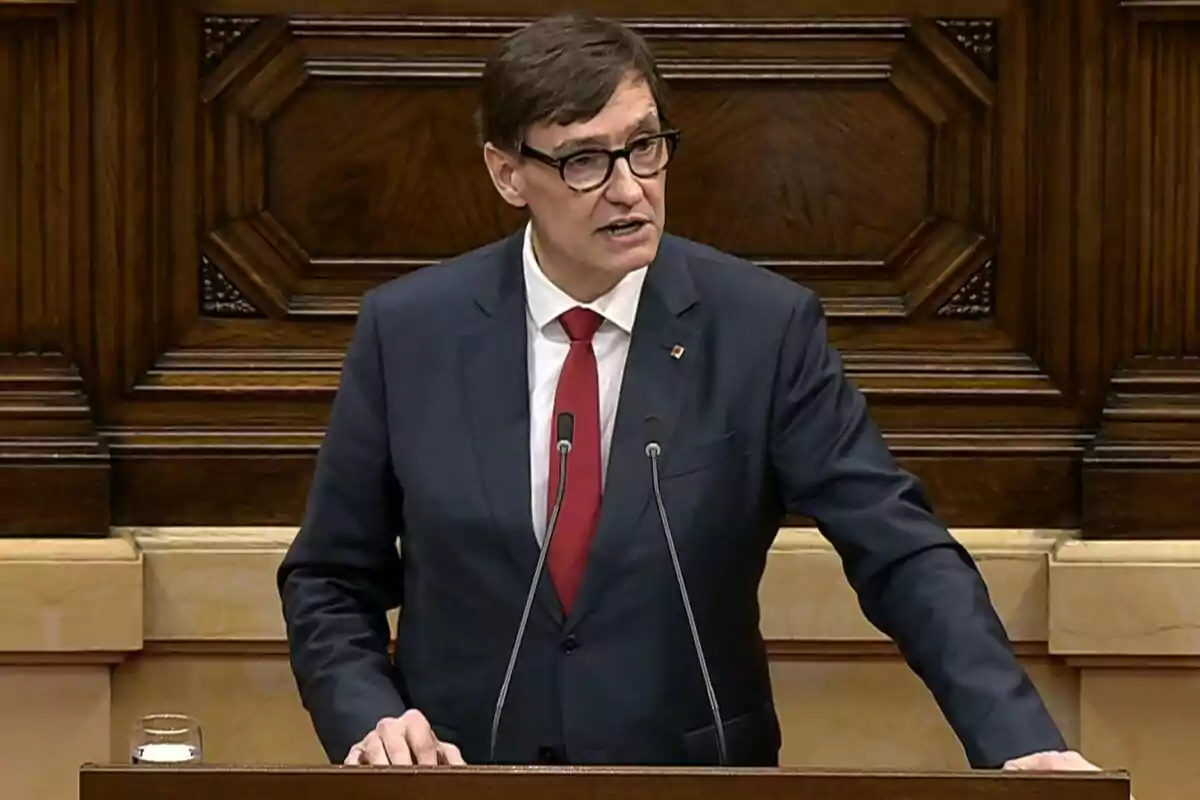 Hombre con gafas y traje oscuro hablando en un atril frente a un fondo de madera tallada