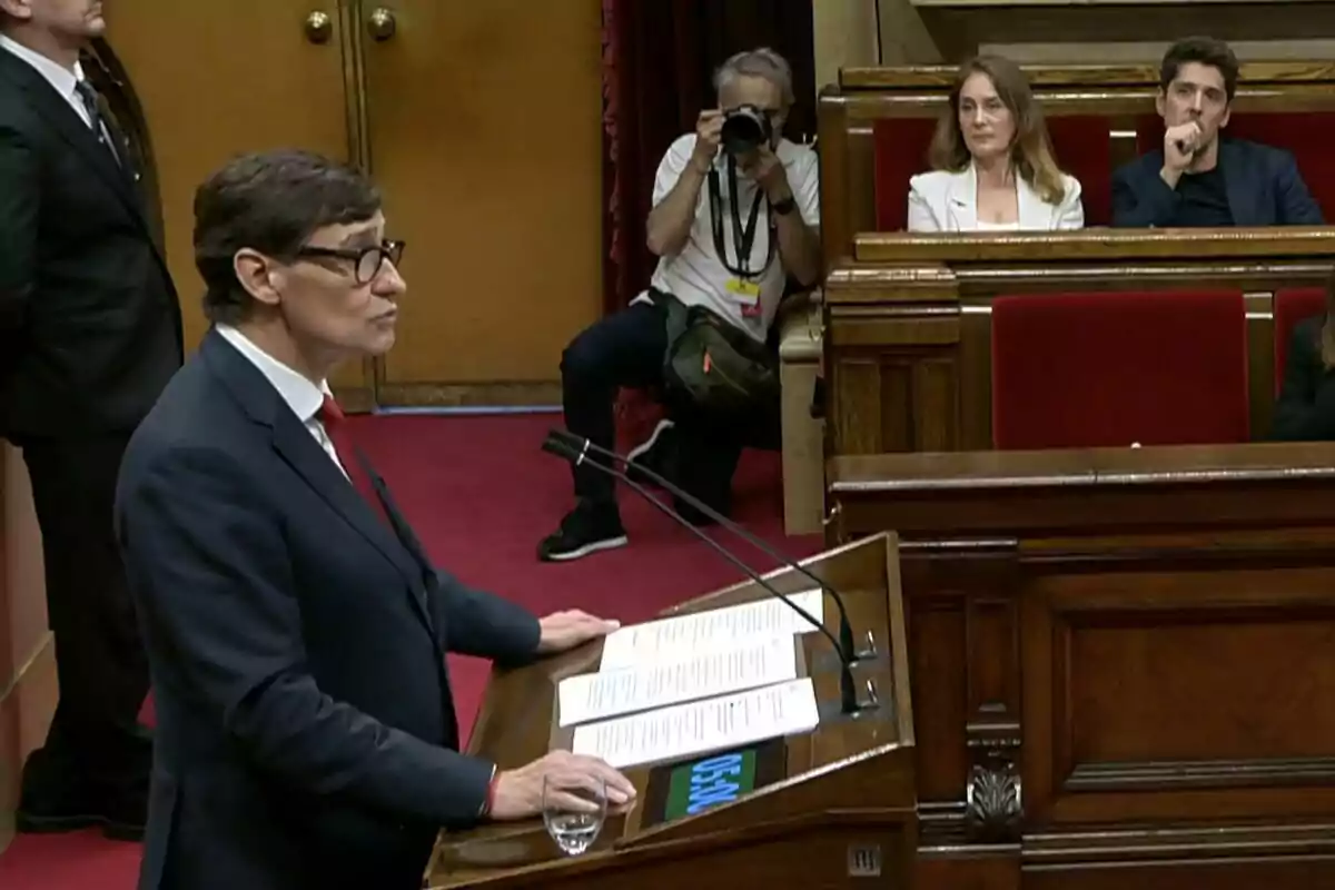 Un hombre con gafas y traje oscuro habla en un atril de madera en una sala parlamentaria mientras un fotógrafo toma fotos y dos personas lo observan desde los asientos.