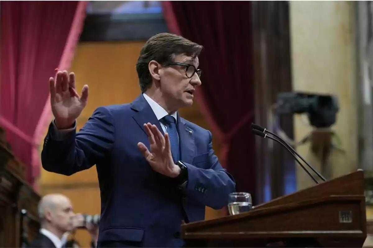 Hombre con gafas y traje azul hablando en un podio en un entorno parlamentario con cortinas rojas de fondo