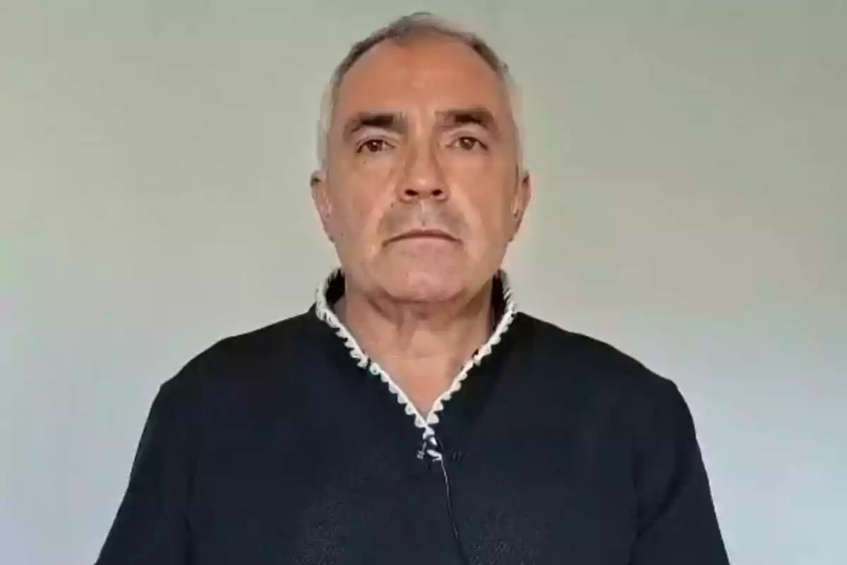 Hombre de cabello canoso y expresión seria vistiendo un suéter oscuro frente a una pared clara