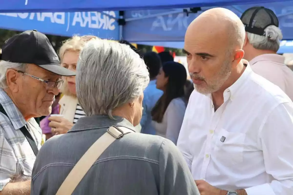 Un hombre calvo con barba y camisa blanca conversa con dos personas mayores en un evento al aire libre bajo una carpa azul rodeados de más asistentes.