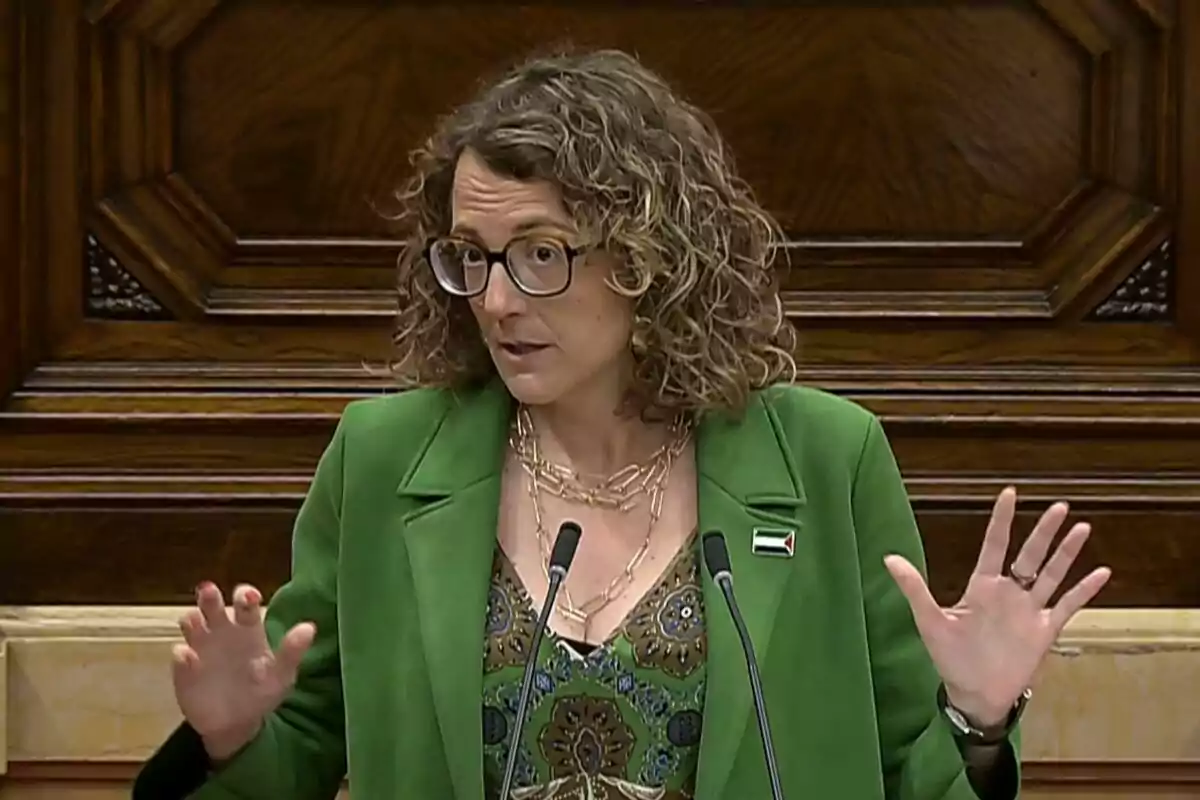 Dona de cabells arrissats i ulleres parlant en un faristol amb jaqueta verda i fons de fusta