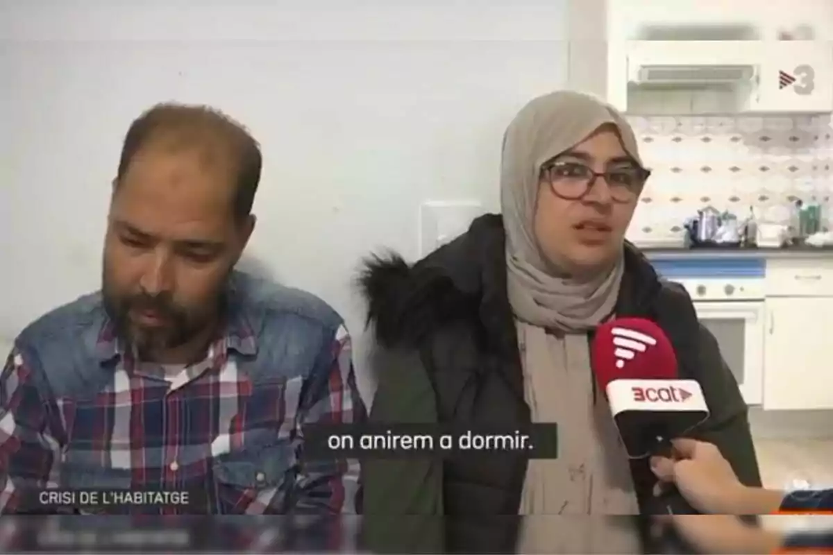 Una mujer con gafas y pañuelo en la cabeza es entrevistada por un canal de televisión mientras un hombre con camisa de cuadros está sentado a su lado en una cocina.