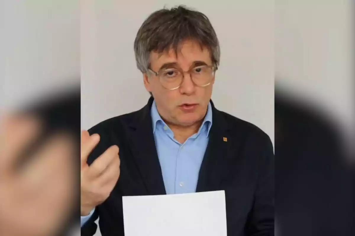 Home de cabells canosos i ulleres amb jaqueta fosca i camisa blava parlant i sostenint un full blanc davant d’un fons clar Home de cabells canosos i ulleres amb jaqueta fosca i camisa blava parlant i sostenint un full blanc davant d’un fons clar