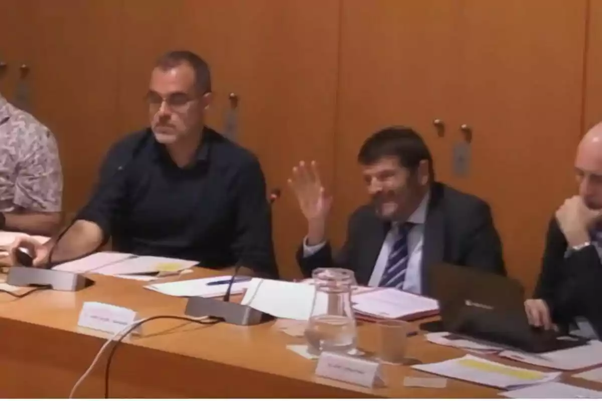 Varios hombres sentados en una mesa de reuniones, uno de ellos levanta la mano mientras los demás observan documentos y computadoras.