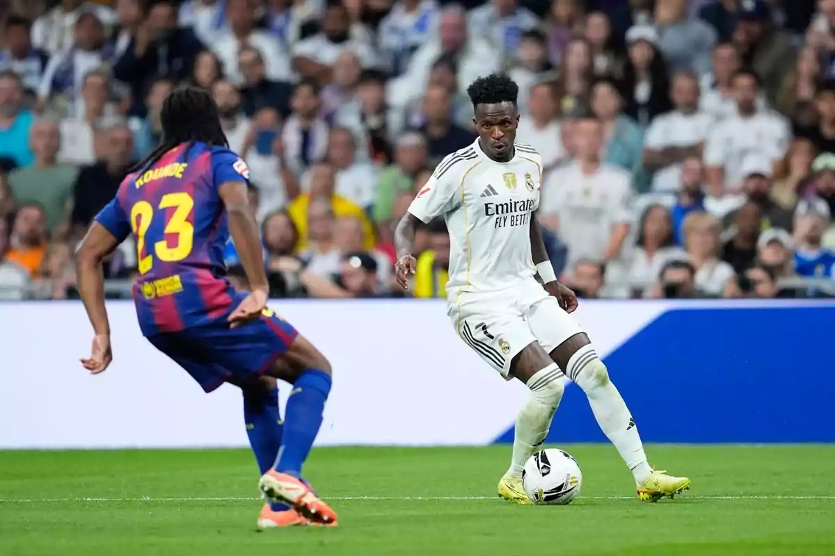 Dos futbolistes dels equips Reial Madrid i Barcelona disputen la pilota durant un partit amb el públic de fons.