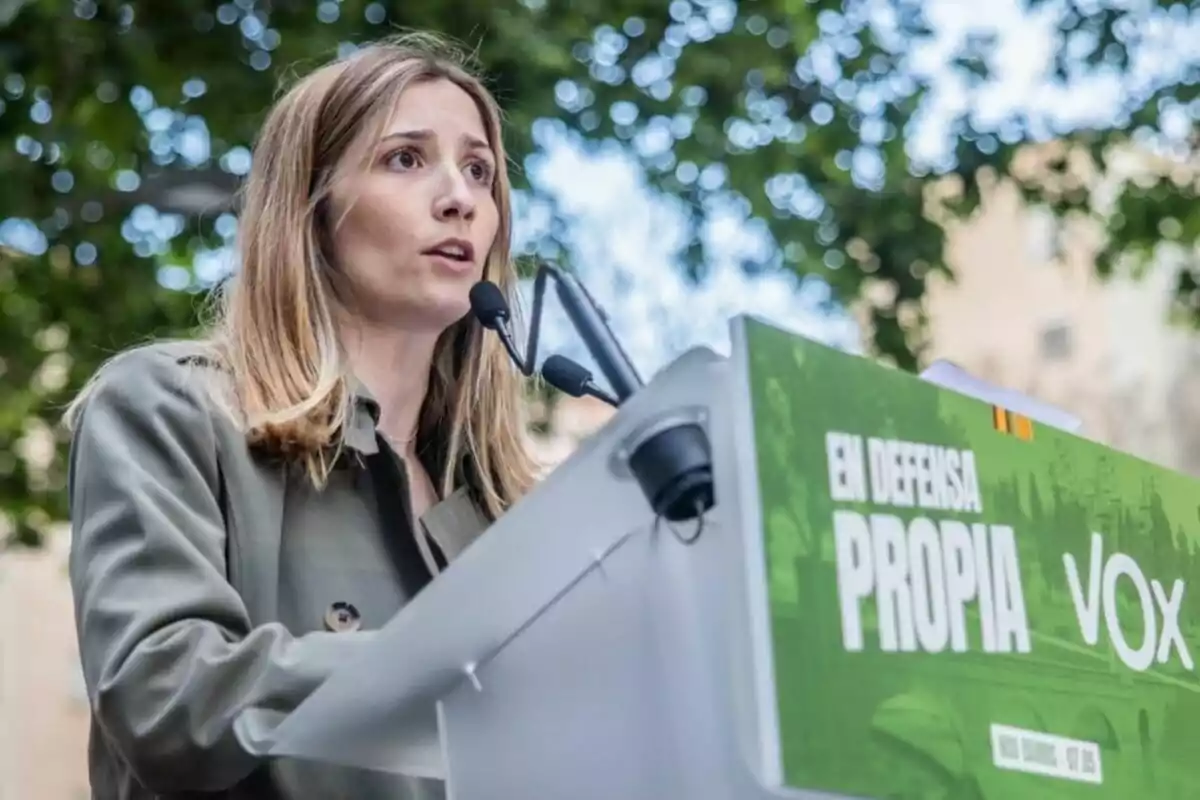 Mujer hablando en un mitin al aire libre frente a un atril con el logo de VOX y un cartel verde que dice En defensa propia