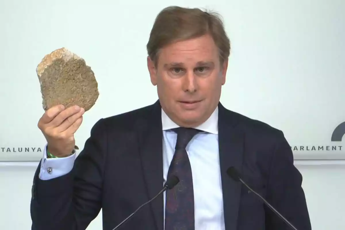 Hombre de traje sosteniendo una piedra grande mientras habla frente a micrófonos en una conferencia de prensa