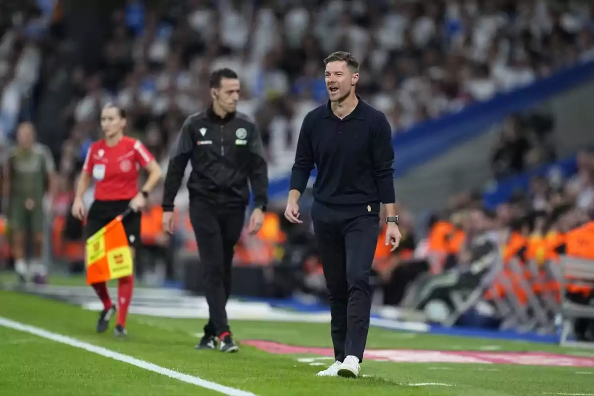Entrenador de futbol donant indicacions a la línia de banda durant un partit amb àrbitres i assistents al fons i públic a les grades Entrenador de futbol donant indicacions a la línia de banda durant un partit amb àrbitres i assistents al fons i públic a les grades